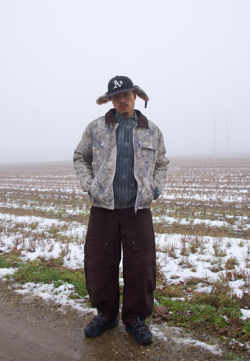 Homme portant une veste camouflage, un pantalon cargo foncé, un pull en tricot, une casquette bordée de fourrure et des baskets, debout sur un champ enneigé sous un ciel brumeux.