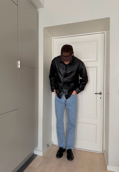 Hombre con chaqueta de cuero negra, vaqueros azul claro, zapatos negros y gorra marrón que dice "El arte de llegar tarde" de pie frente a una puerta blanca.