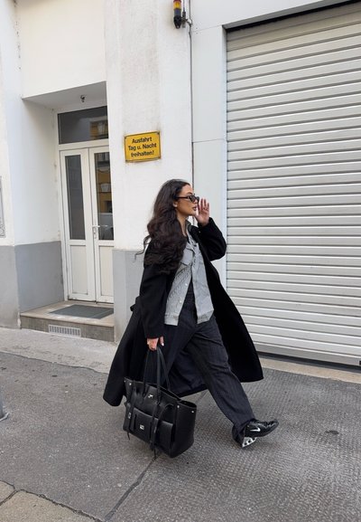 Mujer con abrigo negro y gafas de sol caminando por la acera, llevando un gran bolso negro cerca de un edificio blanco con persiana cerrada y un cartel en alemán.