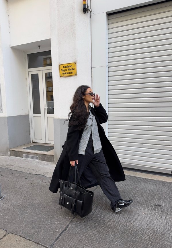 Kobieta w czarnym płaszczu i okularach przeciwsłonecznych idąca chodnikiem, niosąca dużą czarną torebkę obok białego budynku z zamkniętą roletą i niemieckim znakiem.