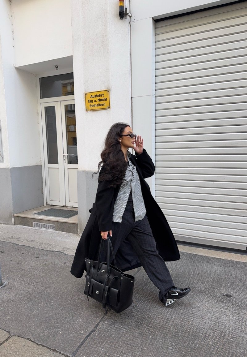 Γυναίκα με μαύρο παλτό και γυαλιά ηλίου περπατά στο πεζοδρόμιο, κρατώντας μεγάλη μαύρη τσάντα κοντά σε λευκό κτίριο με κλειστή περσίδα και γερμανική πινακίδα.