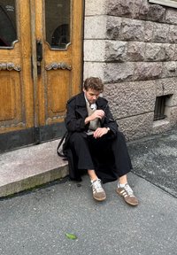 Czarny płaszcz trench, kremowy sweter i ciemne spodnie. Siedzi na kamiennych schodach obok drewnianych drzwi, trzymając okulary. Szare sneakersy z białymi akcentami.