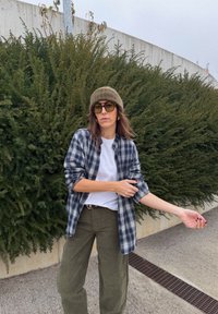 Cămașă în carouri albastre și albe peste un tricou alb, pantaloni verde măsliniu, curea maro, căciulă verde și ochelari de soare rotunzi; fundal verde luxuriant.