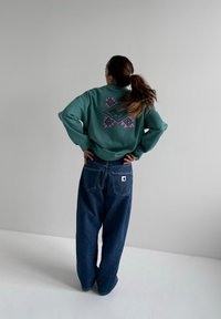 Sweat-shirt vert avec un motif géométrique violet au dos, associé à un jean denim bleu ample avec une étiquette logo sur la poche arrière.