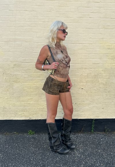Top corto gráfico marrón con un patrón texturizado, combinado con pantalones cortos marrones desgastados. Botas altas negras completan el conjunto.