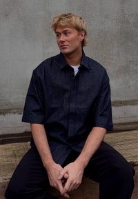 Camicia in denim blu scuro a maniche corte con chiusura a bottoni, dotata di colletto e vestibilità morbida, indossata sopra una maglietta bianca.