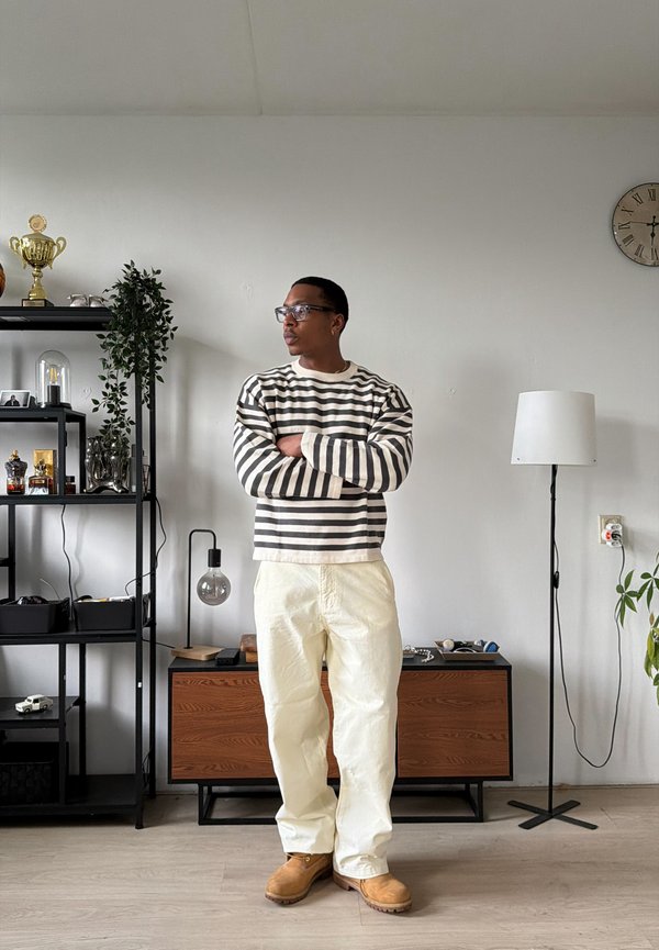 Homme portant des lunettes, chemise longue rayée, pantalon blanc et bottes beige, debout les bras croisés dans une pièce minimaliste.