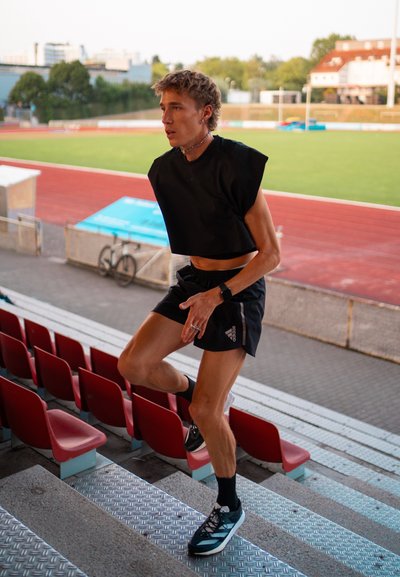 Top corto negro sin mangas, pantalones cortos negros, zapatillas de correr con acentos en color aguamarina. Persona ascendiendo por escaleras de metal en un estadio con asientos rojos.