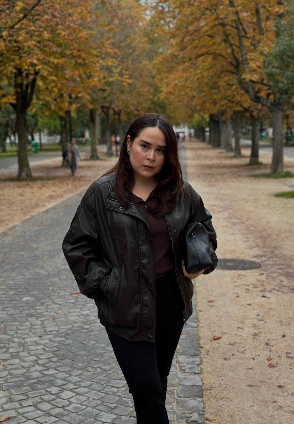 Braune Lederjacke über einem dunklen Oberteil, kombiniert mit schwarzen Hosen. Hält eine schwarze Tasche; im Hintergrund sind herbstliche Bäume und ein Kopfsteinpflasterweg zu sehen.