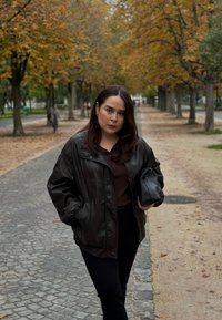 Braune Lederjacke über einem dunklen Oberteil, kombiniert mit schwarzen Hosen. Hält eine schwarze Tasche; im Hintergrund sind herbstliche Bäume und ein Kopfsteinpflasterweg zu sehen.