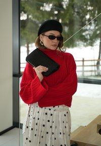 Femme portant un béret noir et des lunettes de soleil, tenant une pochette noire, vêtue d'un pull rouge à torsades et d'une jupe blanche à pois, à l'intérieur près d'une fenêtre.