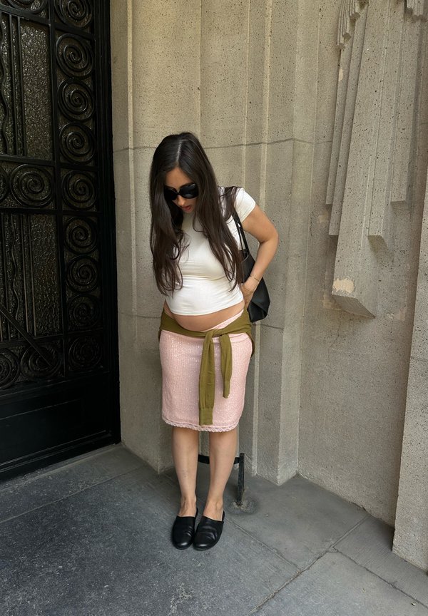 Femme enceinte aux longs cheveux foncés, haut blanc, jupe rose et lunettes de soleil, debout près d'un mur de pierre gris et d'une porte noire ornée.