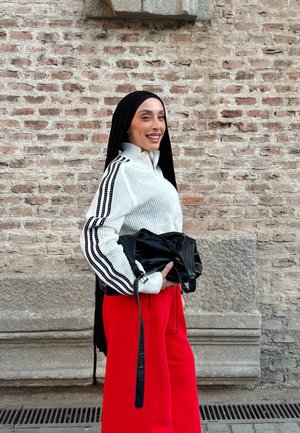 Mujer con hiyab negro, suéter blanco con rayas negras, pantalones rojos, sosteniendo bolso negro, sonriendo, de pie frente a una pared de ladrillos.