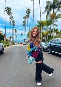 Denim jacket with multicolored graphic prints, paired with black wide-leg pants. Neutral sneakers complete the look against a coastal backdrop.