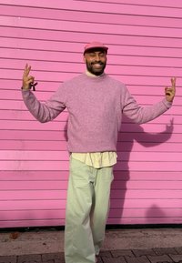 Gaiši rozā džemperis, ar baltu kreklu zem tā un mintzaļas platajiem bikšu galiem. Fonā ir svītraina rozā siena. Persona rāda miera zīmes ar rokām.