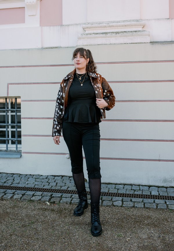 Femme portant une veste marron à motifs, un haut noir, un pantalon, des bottes et des colliers dorés superposés, se tient sur un chemin pavé à côté d’un mur rayé.