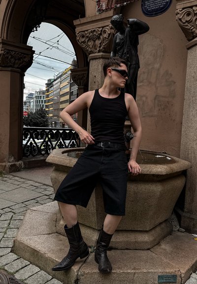 Top sin mangas negro, pantalones cortos negros hasta la rodilla y botas de vaquero marrones. Posando junto a una fuente de piedra con un fondo de estatua.