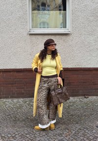 Manteau long jaune, haut côtelé vert clair, pantalon large imprimé léopard marron, béret marron, lunettes de soleil, portant un sac à main marron, debout sur des pavés.