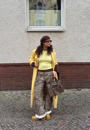 Abrigo largo amarillo, top verde claro acanalado, pantalones anchos marrones con estampado de leopardo, boina marrón y gafas de sol, llevando un bolso marrón, de pie sobre adoquines.