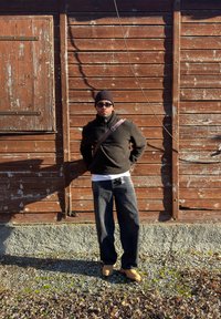 Homme portant un bonnet noir, des lunettes de soleil, un pull sombre, un jean noir et des bottes couleur tan, debout contre un mur en bois usé sous la lumière du soleil.