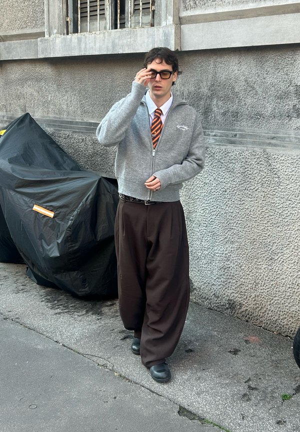 Giovane uomo con occhiali da sole, maglione grigio con zip, cravatta arancione a righe e pantaloni larghi marroni cammina sul marciapiede di una città accanto a una motocicletta coperta.