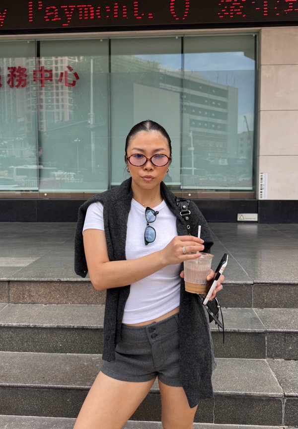 Cardigan sans manches gris par-dessus un t-shirt blanc, associé à un short gris. Tenant une boisson et un smartphone. Des lunettes de soleil rondes complètent le look.