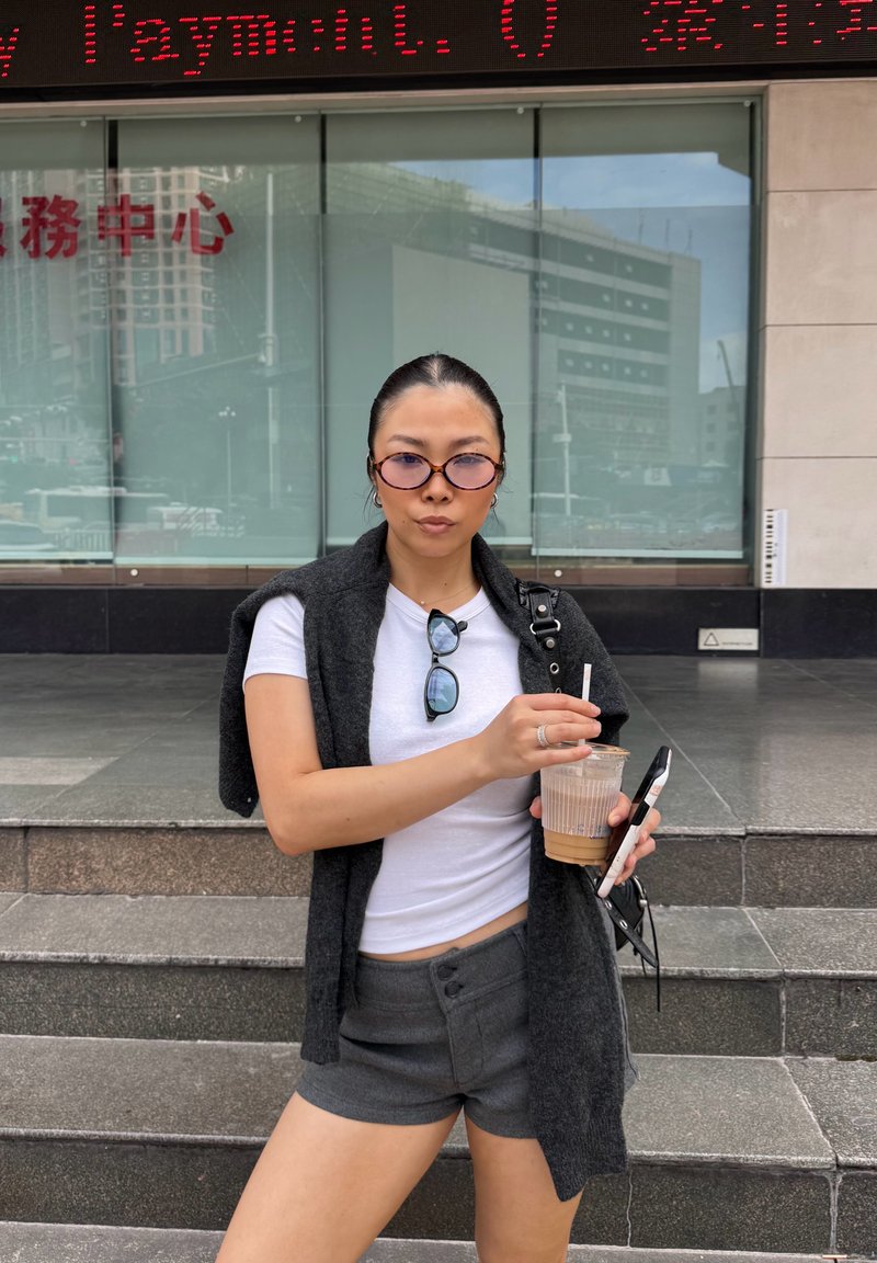 Cardigan grigio senza maniche sopra una maglietta bianca, abbinato a pantaloncini grigi. Tiene in mano una bevanda e uno smartphone. Occhiali da sole rotondi completano il look.