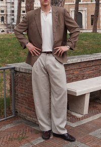Blazer castanho texturizado sobre uma camisa polo branca, combinado com calças em tom bege claro e sapatos castanhos. Pose de pé, com as mãos na cintura.