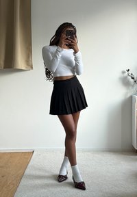 Weisses langärmliges Crop-Top, schwarzer plissierter Rock, weisse Socken und spitze burgunderrote High Heels. Stehend auf einem hellen Teppich, vor einem neutralen Hintergrund.