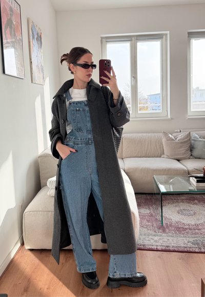 Abrigo largo gris sobre un mono de mezclilla, camisa blanca y botas de plataforma negras. Gafas de sol negras y joyería dorada acentúan el look.