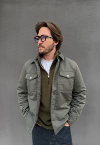 Mees pruunide juuste ja habemega, kandes suuri prille, oliivroheline jakk fliisist kampsuni peal, käed taskutes, seisab halli seina ees.