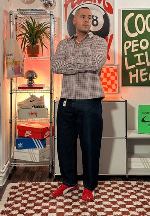 Camisa de cuadros gris y blanca, jeans azul oscuro, zapatillas rojas. El fondo presenta estanterías con zapatillas y decoración, incluyendo una planta en una maceta.