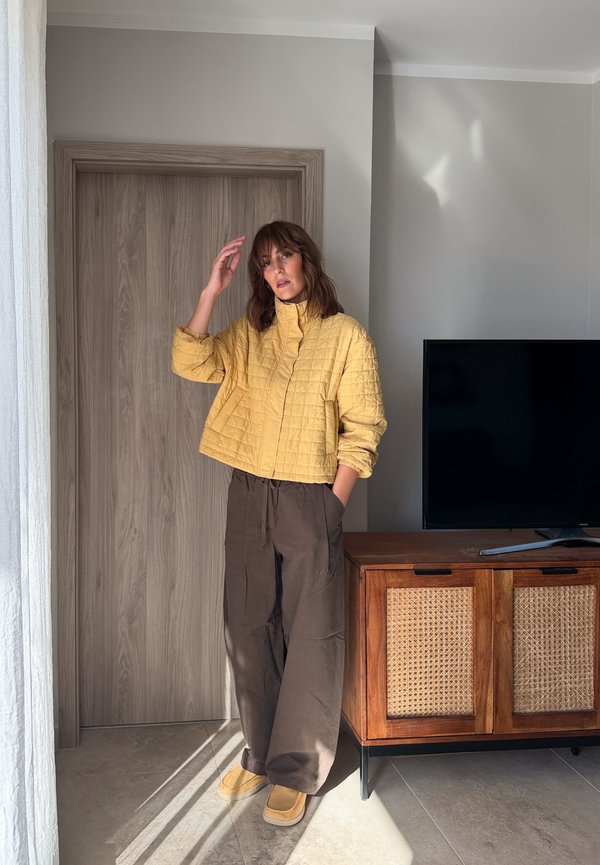 Une femme pose près d’une porte en bois, vêtue d’une veste matelassée jaune, d’un pantalon brun et de chaussures jaunes. À proximité, un téléviseur est posé sur un meuble.