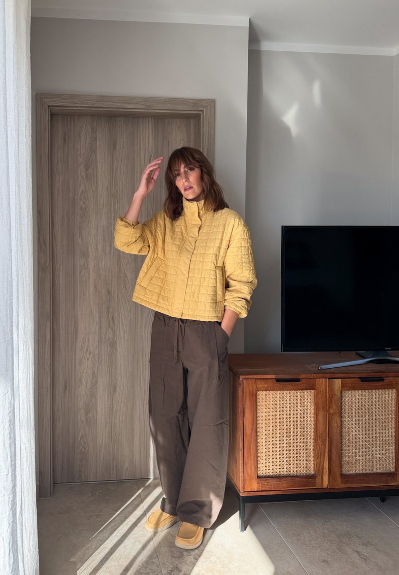 Une femme pose près d’une porte en bois, vêtue d’une veste matelassée jaune, d’un pantalon brun et de chaussures jaunes. À proximité, un téléviseur est posé sur un meuble.