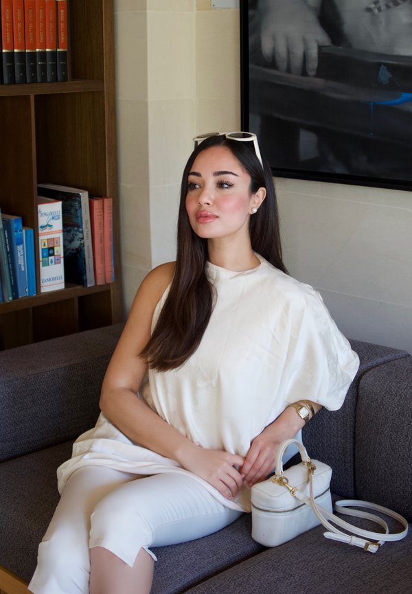 Woman with long dark hair and white outfit sitting on a gray sofa holding a white handbag, with bookshelves and a black-and-white photo behind her.