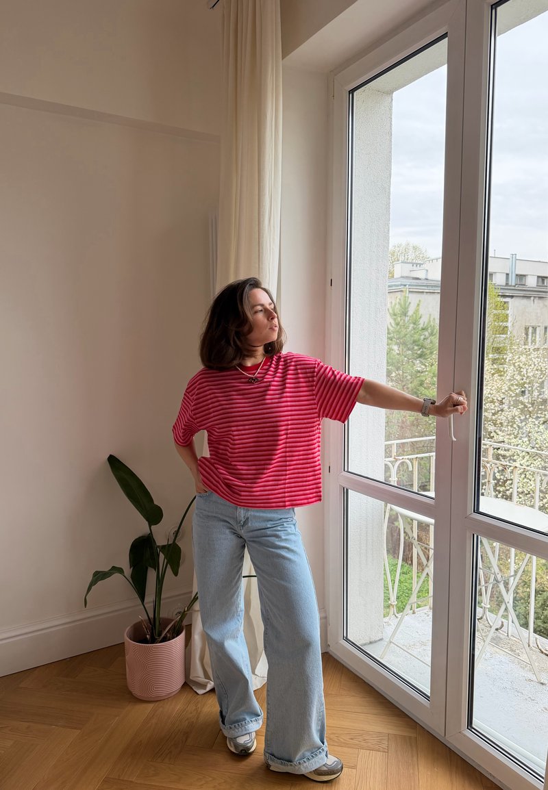 Vrouw in rood gestreept hemd en wijde jeans opent glazen deur, staand op houten vloer nabij een kamerplant en lichte gordijnen.