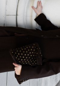 Personne portant un manteau marron foncé tenant un sac à main marron foncé avec des œillets dorés devant un mur texturé blanc.