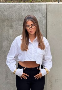 Camisa blanca recortada con un bolsillo y mangas extragrandes, combinada con pantalones negros ajustados y detalles de pulseras doradas. Cabello liso.