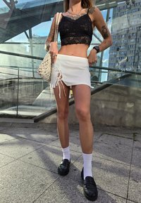 Black lace crop top, white mini skirt with side ties, black loafers, white socks; natural surroundings and architecture in the background.