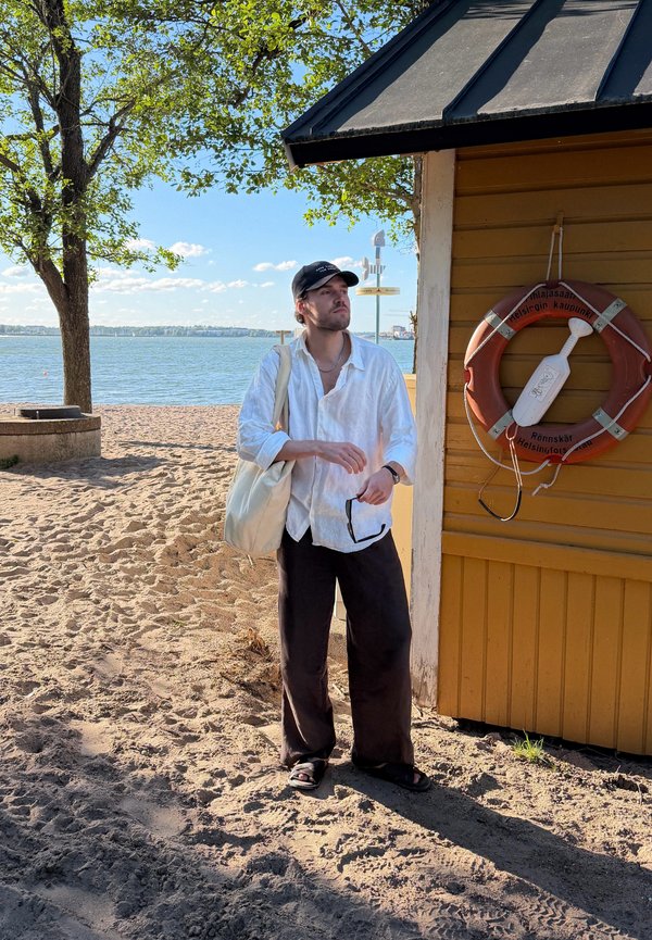 Hombre con camisa blanca y gorra negra está de pie en una playa de arena cerca de un edificio de madera amarillo con un salvavidas, mirando hacia el agua.