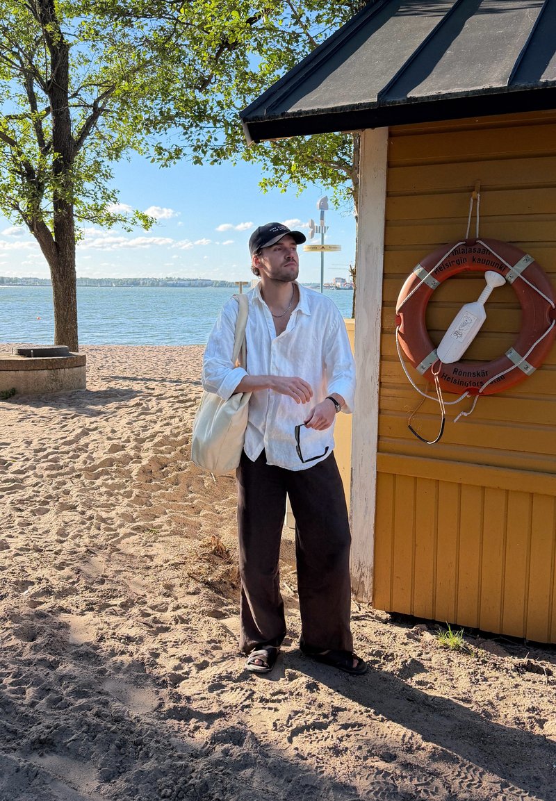 Homme en chemise blanche et casquette noire debout sur une plage de sable près d'un bâtiment en bois jaune avec une bouée, regardant vers l'eau.