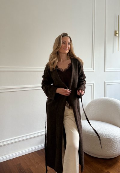 Mujer con largo cabello rubio vistiendo un abrigo marrón oscuro sobre una blusa negra y pantalones crema, de pie junto a un sillón blanco texturizado en una habitación luminosa.