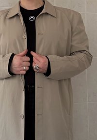 Personne boutonnant un manteau beige par-dessus une chemise noire ornée d'un petit logo circulaire blanc, portant deux bagues argentées aux doigts.