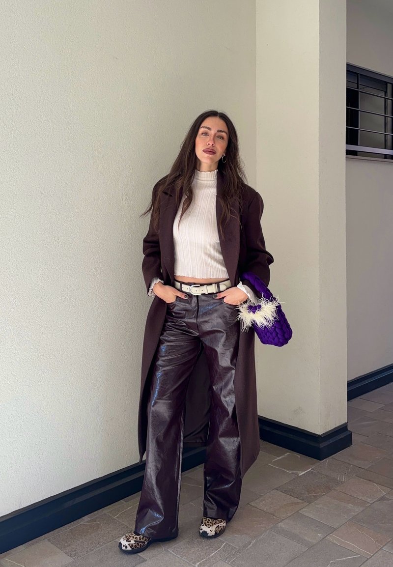 Donna con cappotto lungo scuro, dolcevita bianco a coste, pantaloni scuri lucidi, scarpe con stampa leopardata e borsa viola con piume bianche.