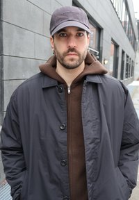 Άνδρας με γένια και σκουλαρίκια που φορά γκρι καπέλο, καφέ φούτερ και σκούρο σακάκι, στέκεται έξω κοντά σε γκρίζους τοίχους κτιρίου.