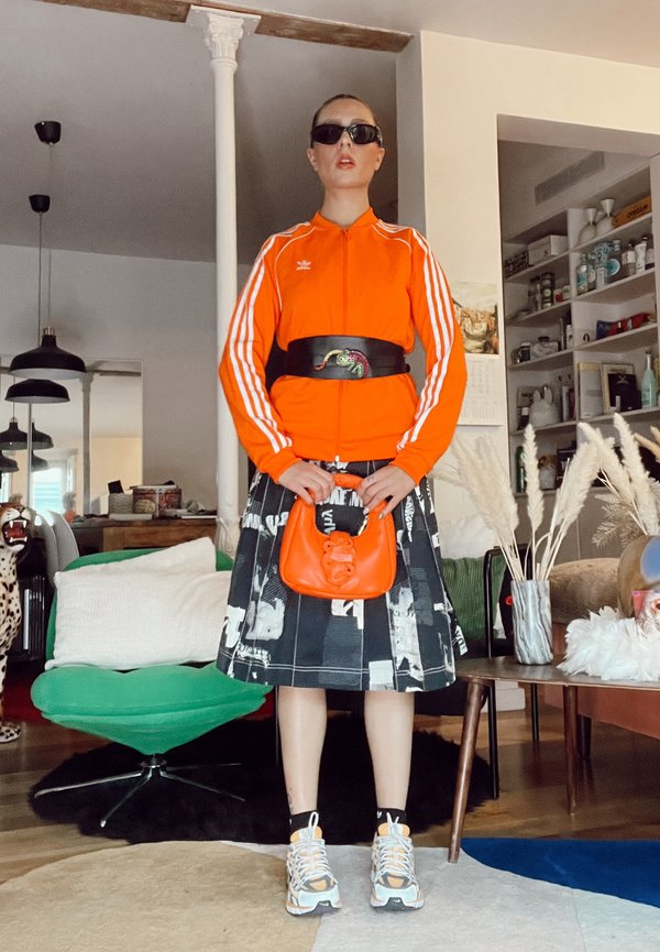 Oranžinė Adidas užtrauktuku užsegama striukė su baltais dryžiais, juoda sijonas su baltais spaudiniais, oranžinė rankinė ir sportbačiai su geltonais akcentais.