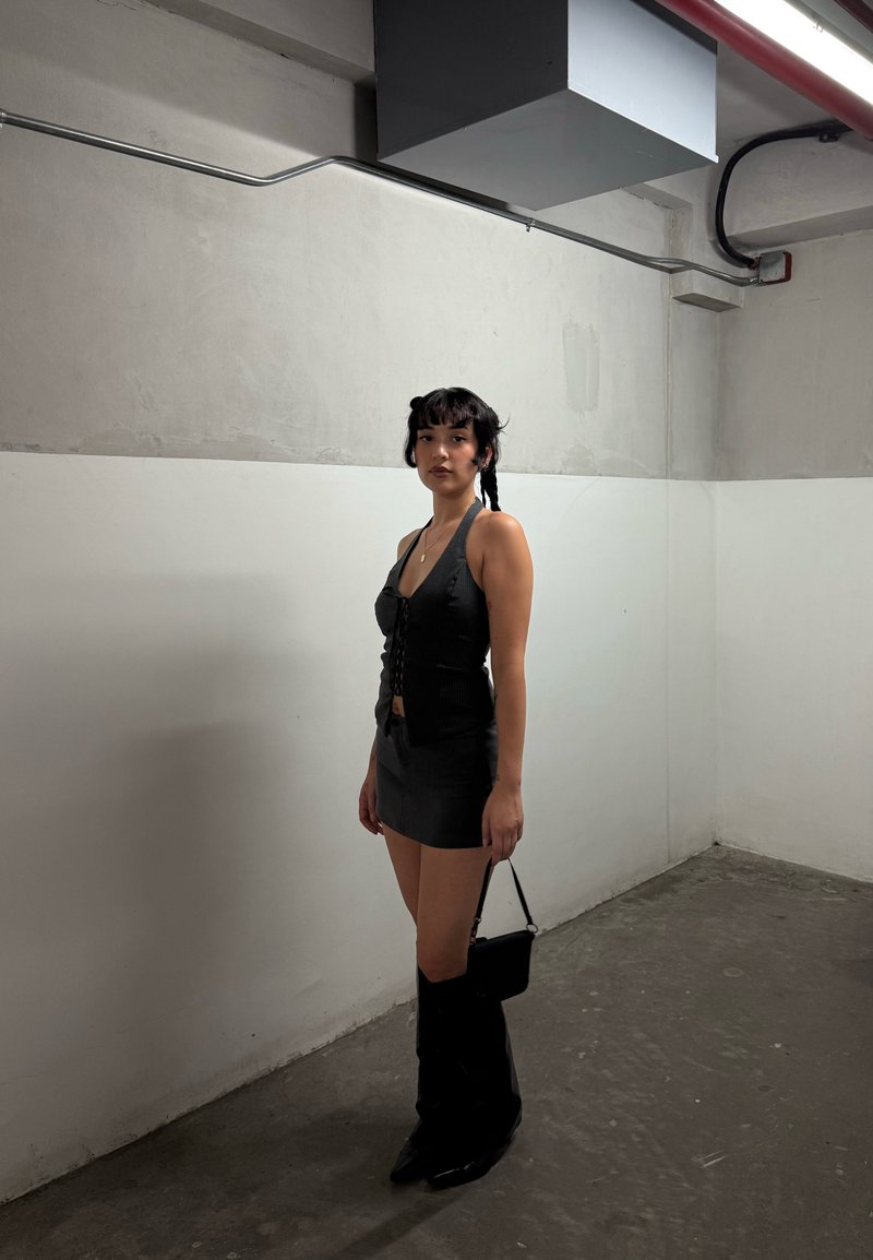 Mulher com cabelo trançado vestindo uma blusa preta sem mangas, saia mini, botas até ao joelho, segurando uma pequena bolsa preta em um quarto de concreto simples.