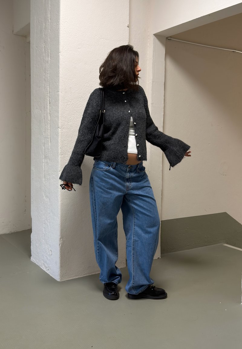 Frau mit schulterlangem dunklem Haar, die eine dunkelgraue Strickjacke, ein weißes Oberteil, lockere blaue Jeans, schwarze Schuhe trägt und eine Brille sowie eine schwarze Tasche in der Hand hält.