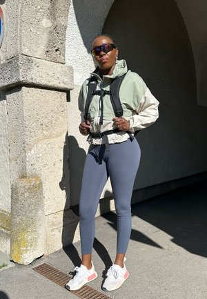 Rompeviento ligero bicolor en verde y crema, leggings grises y zapatillas blancas con acentos rosas, combinados con una mochila y gafas de sol.