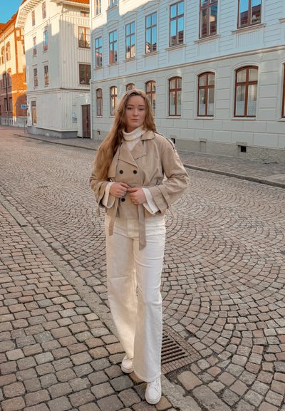 Mujer joven con el pelo largo, que lleva una chaqueta beige y pantalones blancos, de pie en una calle empedrada bordeada de edificios blancos y de ladrillo.